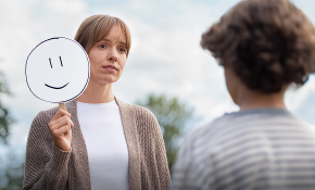 Depressed woman eating breakfast with family while holding a smiley face sign.